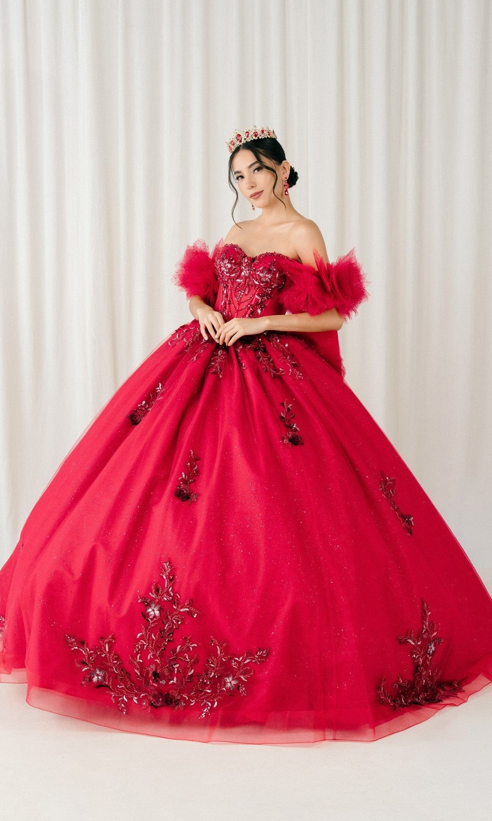 A woman wearing the Quinceanera Dress 8380J By Cinderella Couture in burgundy, featuring floral embellishments and a corset bodice, stands before a white curtain backdrop with a crown.